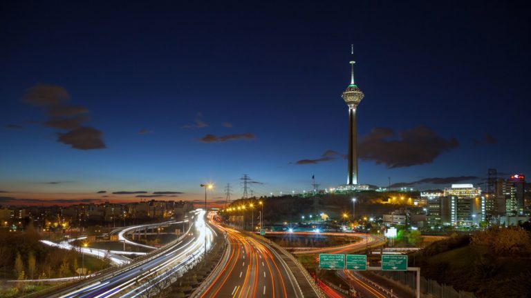 Night view of a city with a tall illuminated tower and busy highway traffic.