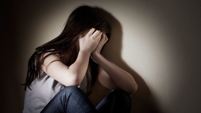 A woman sits alone, covering her face in distress.