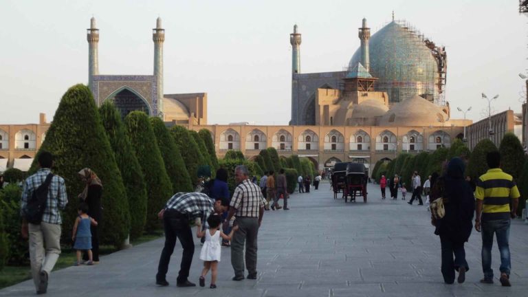 People strolling in a historic square with domed mosque towers.