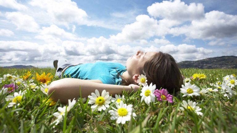 A young woman lies peacefully in a flower-filled meadow under a partly cloudy sky.