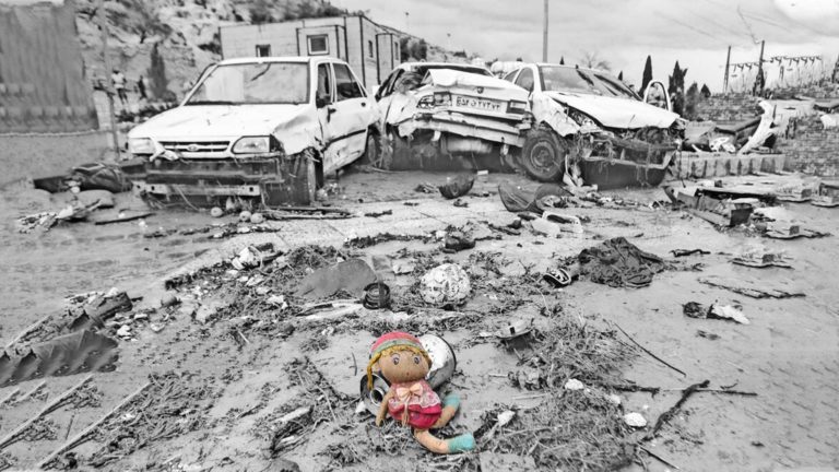 A child sits alone in muddy debris near wrecked vehicles after a disaster.