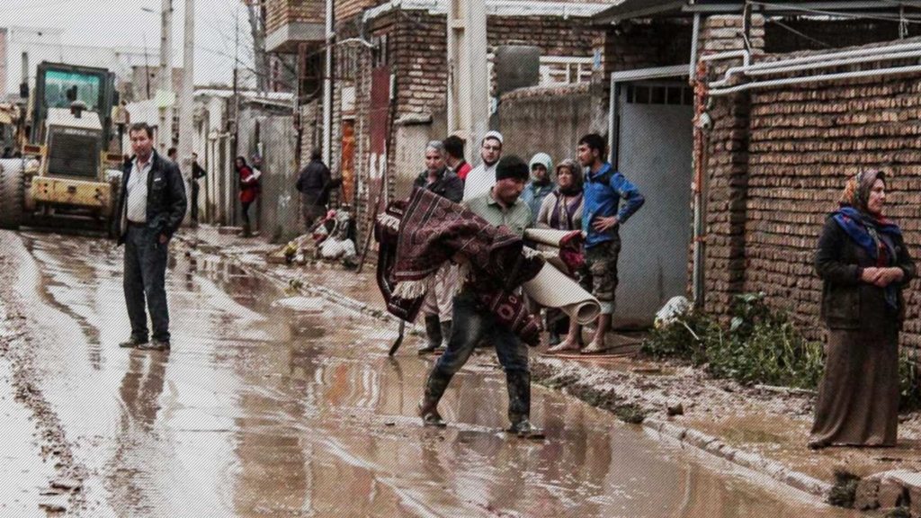 Man carries a child through a flooded, muddy street in a poor neighborhood.