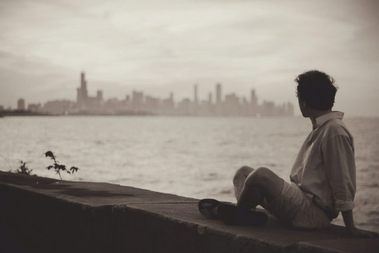 Person sitting by the water, overlooking a distant city skyline in sepia tone.