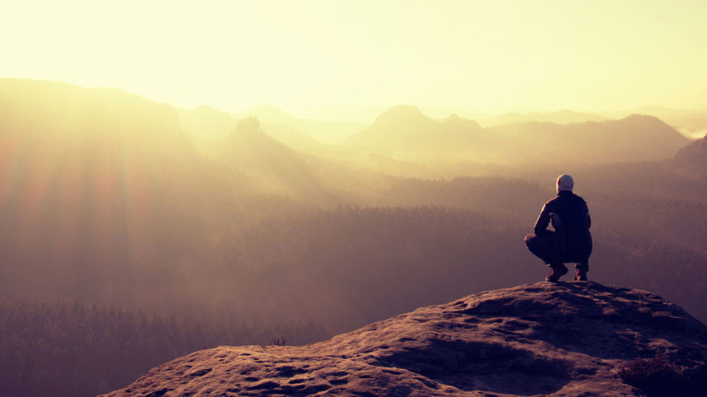 Person sitting on cliff edge overlooking hazy mountains at sunrise.