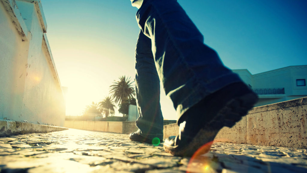 Close-up of a person walking on a sunlit street with palm trees in the background.