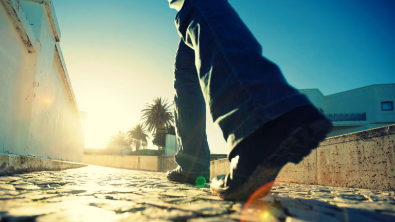 Close-up of a person walking on a sunlit street with palm trees in the background.